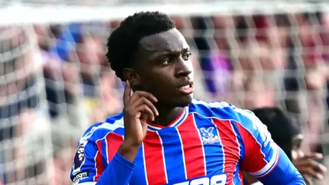 Eddie Nketiah seen from chest up in red and blue vertical striped Crystal Palace shirt running away in celebration, putting his right index finger up to his ear as if to show he is listening. In the background a white net is visible through which a blurred crowd can be seen