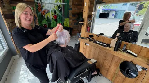 Kevin Shoesmith/BBC News A woman with long blonde hair cuts an older woman's hair. The stylist is smiling for the camera.