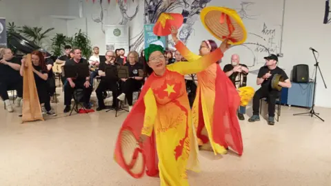 Women dance in red and yellow dresses as musicians pay behind them