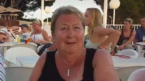 McGrath Family June McGrath pictured looking at the camera in a sun holiday setting. She has short, blonde hair and is wearing a black vest top and a silver pendant necklace. The pensioner is sitting in an outdoor bar or cafe which has white plastic tabled and chairs. Behind her are other customers in summer clothing, trees and thatched parosols. 