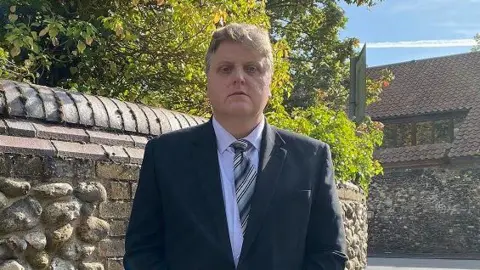 Gregory Hill, dressed in a suit, standing in front of a flint wall his hands clasped in front of him