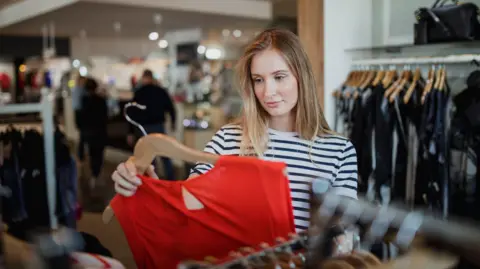 A young woman in stripy blue and white top looks at a red dress in a shop 