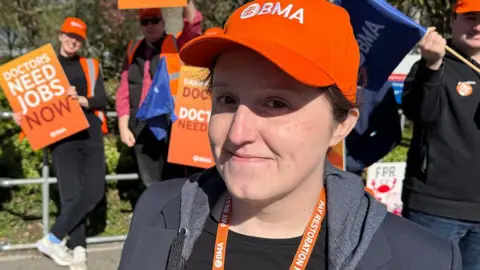 Dr Becky Lavelle is wearing an orange BMA cap and a navy blazer over a dark hooded top. She is standing on a picket line during a street protest on a sunny day smiling slightly, while other doctors behind her hold orange placards that read: "DOCTORS NEED JOBS NOW".