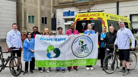Two cyclists standing for a photo outside a building with people cheering them on stood between them. An ambulance is behind them.