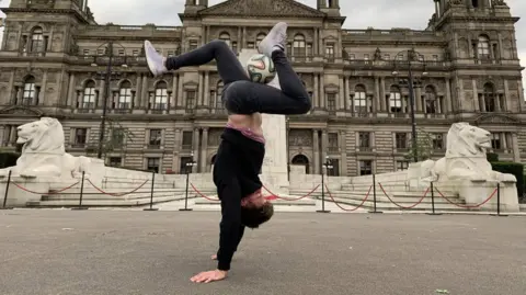 Ross Brown COPYRIGHT PERMISSION TBC*** A man in jeans, trainers and a black jumper is doing a handstand in front of a large stately home building. His legs are bent at right angles and a football is being balanced on one of his legs.
