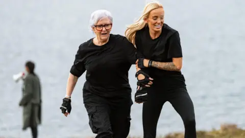 BBC/Studio Lambert Linda, who has purple/grey hair and glasses, is running up a lake shore. She is being helped by Leanne, another contestant, who is tanned with long blonde hair and a tattooed forearm. Both are dressed in all black with gloves. They are wearing expressions of stress.