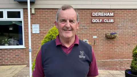 Tom Williams/BBC Stuart Goodman, wearing a purple shirt and navy jumper, is standing in front of the clubhouse, with a sign on the wall behind that reads Dereham Golf Club.