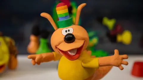 Jason lock Prop of Chorlton the Happiness Dragon with a rainbow striped hat and yellow top with hands stretched out.