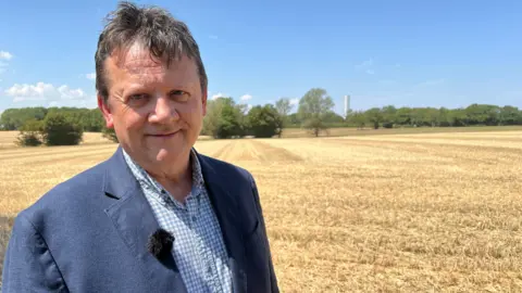 Mousumi Bakshi/BBC A man standing in a large field with what looks like a white cooling tower in the very background