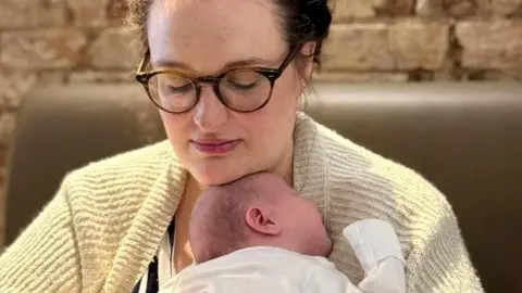Charlie holds her baby June on her chest, while wearing a yellow cardigan. The baby is wearing a white baby suit