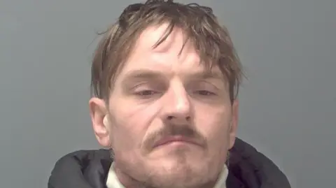 A mugshot of a man wearing a hooded jacket over a white rollneck jumper. He is standing in front of a grey background and looking into the camera.