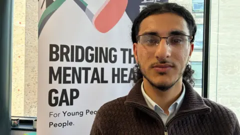 Rushid has long black hair and a beard and is wearing glasses, a blue and white shirt and a brown zipped top. He is standing in front of a white banner which says "bridging the mental health gap" in black letters.