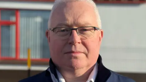 Dermot O'Brien is wearing black glasses. He has a white shirt and navy jacket on. He has short grey hair. The background is blurry. 