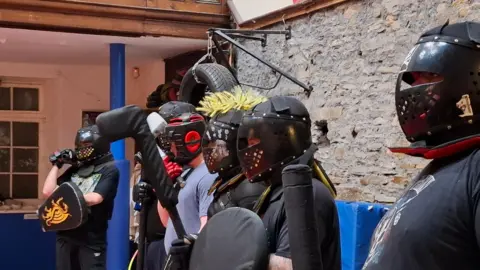 Medieval Combat Wales Line of people standing indoors wearing black protective fencing-style helmets with perforated faceplates and padded armour. Some hold round shields and foam-covered training weapons. One helmet has a bright yellow crest attached on top. The setting appears to be a training hall with a stone wall, wooden panelling, and a tyre hanging from a metal bracket overhead.