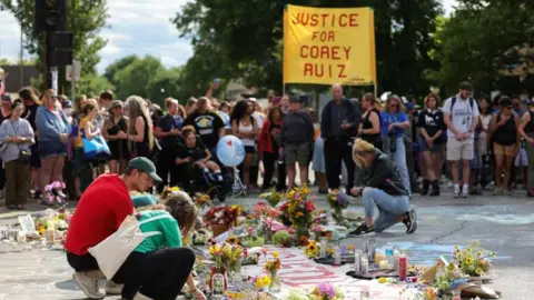 Reuters People bend over to read notes and flowers left in honour of Ruiz. In the background a group of protesters gather and one holds a bright yellow sign that reads "Justice for Corey Ruiz". 