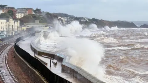 The picture shows huge waves crashing over a sea wall during rough weather. Brown, churning water is surging in from the right, and the force of the waves is sending spray high into the air. A railway line runs along the left side of the image, curving around the bay, with wet tracks glistening from the spray. Buildings sit close to the shoreline, and rows of houses climb up the hillside in the distance. The sky is grey and overcast, and the whole scene looks stormy and chaotic, with the sea battering the coast.