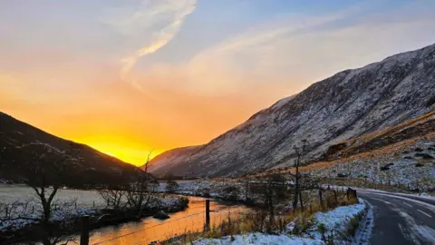 Anna Campbell A slightly snowy road with a hill on one side and a river on the other. At the end is the sunrise which is yellow and orange.