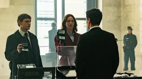 Disney via Getty Images O personagem de Evan Peters usa camisa, gravata e jaqueta e segura sua carteira de identidade do FBI e o personagem de Rebecca Hall usa jaqueta vermelha, camisa e também segura uma carteira de identidade do FBI na frente de um homem, de quem só vemos as costas.