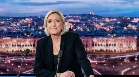 Marine Le Pen sat in a tv studio with a background of Paris wearing a black suit