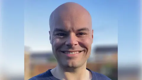 Ben smiles at the camera in a close-up picture against a blue background. 