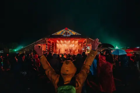 Recompose A brightly lit stage viewed from a distance with a fan standing close to the camera with their hands up in the air