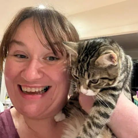Marie Fox Marie Fox holding up Daisy the cat. Marie is smiling at the camera. The cat has striped fur in shades of brown, black, and grey as well as white paws and white on her face