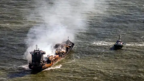 PA Media Smoke billows from a rust-coloured tanker ship floating in the sea. A tugboat floats nearby.