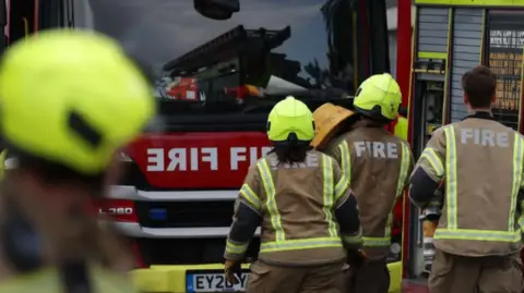 Three firefighters walk towards a fire engine with another crew member blurred in the foreground. They wear brown uniforms with fluorescent strips, with 'fire' printed on the uniforms in big white letters.