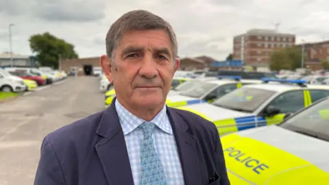 A man in a dark suit and blue tie looks at camera with a row of Wiltshire Police cars in the background, and a police station in the distance.