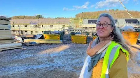 Joanna wears an orange, green and blue scarf and high-viz jacket as she stands side-on, turned toward the camera. In the background is a housing site, with planks fo wood, skips a crane and to the left a row of homes under construction which are covered with scaffolding.