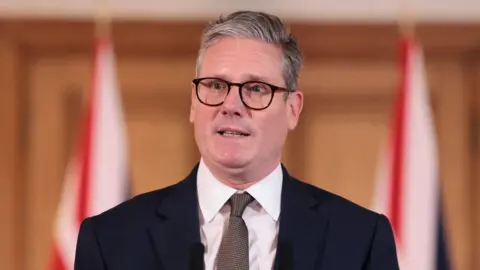 Reuters Sir Keir Starmer standing at a microphone wearing glasses, a navy blazer, white shirt and grey tie