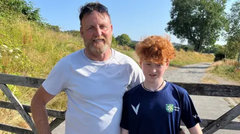 Micahel Thornborough and Rowan stand in front of a wooden farm gate. Michael has short grey hair and sunglasses on top of his head. He is wearing a white t-shirt. Rowan has curly ginger hair and wears a blue t-shirt.