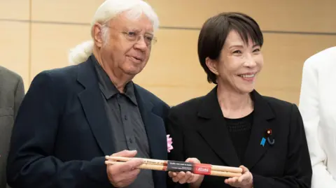 Japanese Prime Minister Sanae Takaichi poses with Ian Paice. They hold a set of signed drumsticks between them. Takaichi has short black hair and wears a black blazer over a black top. Paice has long white hair, tied back, and wears glasses, a black blazer and a grey buttoned shirt.