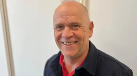 Deputy Marc Lainé - A bald man smiling at the camera, wearing a black jacket and a red polo shirt. 