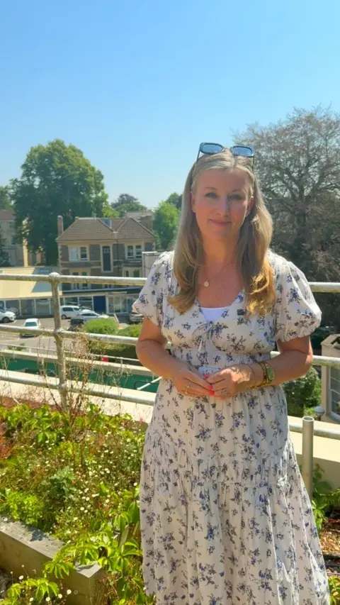 Sara Thornton is standing in front of a border containing green plants, some of which have small white flowers. There are railings at the back of the border. There are trees, a house and some cars in the background. Sara has long, fair hair. She is wearing a white dress with a green and blue floral pattern. She has sunglasses balanced on top of her head. She has pink nails and is wearing a gold necklace.