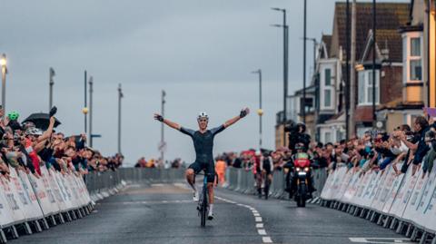 A cyclist holding his hands up in victory with people lining the street in support 