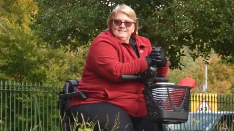 Sue Myerscough seorang wanita dengan mantel bulu merah sedang duduk di atas skuter mobilitas. Dia memiliki panjang bahu, rambut coklat muda, dan mengenakan kacamata hitam. Dia berada di jalan setapak di tengah taman dan dikelilingi oleh pohon dan tanaman hijau.