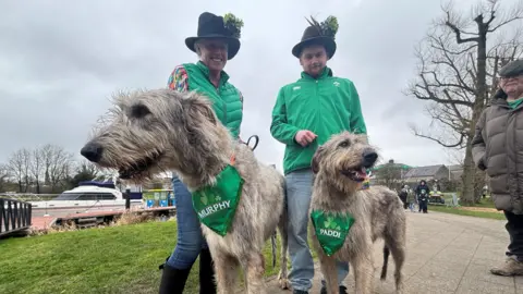 Two Irish wolfhounds are wearing green bibs around their necks. Both have shamrock designs on them. One says Murphy, the other says Paddi. Their owners are wearing hats with shamrock attached and green jackets. 