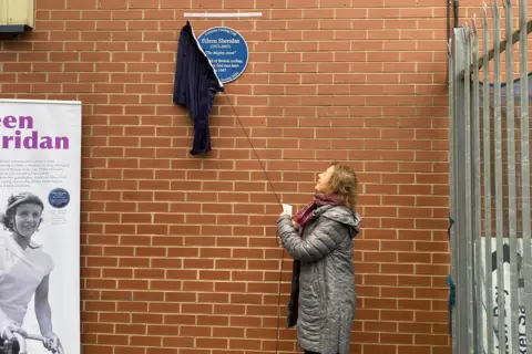 Louise Sheridan wearing a long silver puffer coat unveiling a blue plaque on a red brick wall