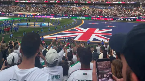 Elliot Family A view of a packed football stadium through a group of fans towards a massive New Zealand flag on the pitch
