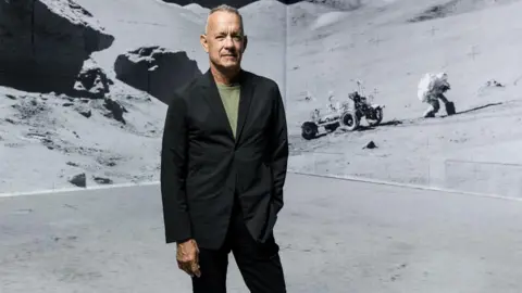 Justin Sutcliffe Tom Hanks in black suit and green T-shirt stands in front of digitally remastered shot of an astronaut and the Moon Rover on the surface of the moon.