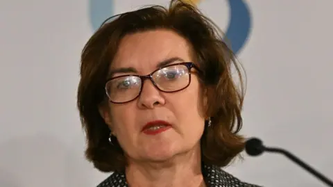 Close up image of Eluned Morgan talking at a government event. She is talking whilst looking out at the audience. She has brown short hair and is wearing glasses. 