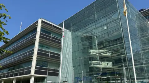 Handout There is a glass building which has Suffolk County Council written in blue on the glass building. There are three flag poles two in the foreground. They are flying the Suffolk flag and St George's Cross. On the top of the building there is also a flag pole which has the Union Jack flag flying on it. 