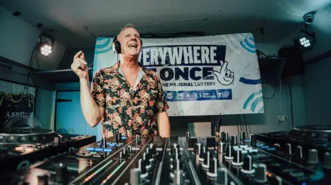 DJ Fatboy Slim stands behind a mixing console inside a low-ceiling venue, wearing headphones and a patterned shirt, with turntables, sliders and dials in the foreground and a large Everywhere At Once banner on the back wall under stage lighting.