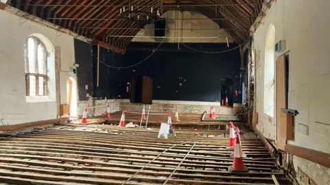 PA Media Looking towards the back of the theatre where there are cones on the floor boards. Two doors are at the end of the building, and the ceiling is arched with wooden beams.