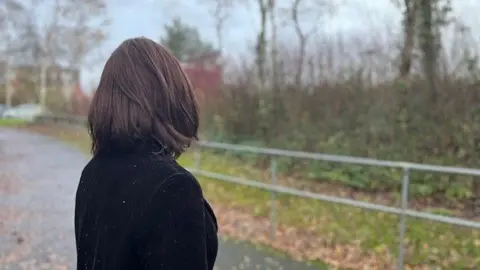 An anonymous woman in a black coat is looking away from the camera and standing on a footpath with shrubbery on either side,