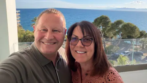 Jason Walker Jason and Michelle take a selfie on the balcony of a hotel. He wears a taupe polo shirt and she wears a salmon woven top. He has dark hair and wears varifocal glasses. In the background there are a few trees and a clear view of the sea.