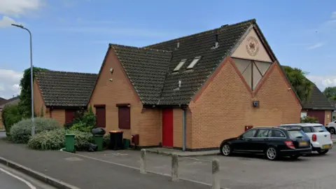 Google The outside of a doctors surgery, which is a brown brick building, with cars parked outside.