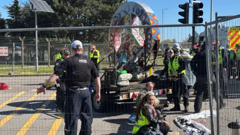 Luke Deal/BBC Demonstrators sitting on the floor and attached to a vehicle outside the entrance to RAF Lakenheath. Police officers are also on the scene. The image is taken from one side of some metal fencing. 