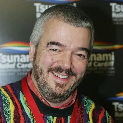 Getty Images Paul Sargent, with brown hair, black beard and colorful jumper, smiles for the camera at a 2005 concert